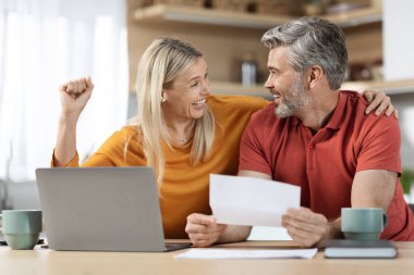 Emotional spouses sitting at table full of papers, laptop, coffee mugs, reading letter, smiling and raising hand up, got good news, kitchen interior. Economy, family budget, mortgage concept