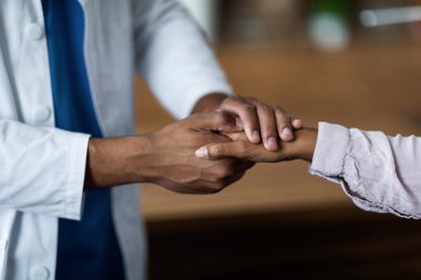 Unrecognizable dark-skinned man doctor supporting female patient, holding her hand, closeup shot, modern clinic background, copy space. Doctor-patient relationship concept