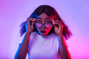 Unbelievable offer. Pretty young black woman touching glasses, looking at camera in shock, opening mouth in surprise, neon light. Amazed millennial African American lady cannot believe huge sale