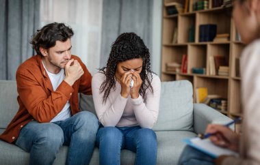 Despaired sad young european man calms black crying woman at meeting with psychologist in office clinic interior. Quarrel and grief, family psychological therapy, depression and relationships problems