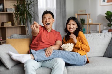 Happy asian middle aged man and young woman in homewear sitting on sofa in living room, cuddling, eating popcorn and laughing, watching comedy movie together at home, pointing at screen, copy space