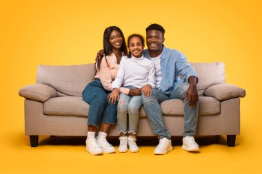Happy black man, woman and girl sitting on couch and embracing over yellow studio wall. Excited daughter posing with her mom and dad smiling at camera. Healthy family relationship concept