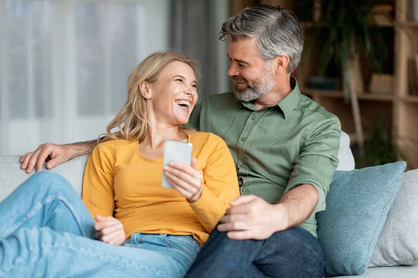 Portrait Of Cheerful Middle Aged Couple Resting With Smartphone On Couch, Happy Mature Wife And Husband Relaxing On Sofa With Mobile Phone, Having Fun Together In Living Room, Closeup Shot