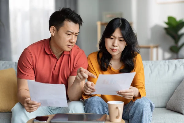 Confused asian spouses checking bills, sitting on couch, reading documents, unhappy chinese man and woman holding papers, counting monthly spendings, copy space. Family budget concept