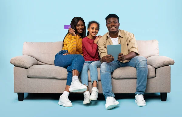 Happy young african american family of three using digital tablet and credit card for online shopping, purchasing goods from internet while sitting on couch over blue background
