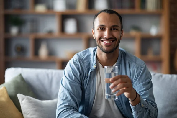 Young Handsome Black Guy Drinking Mineral Water From Glass At Home, Positive African American Man Enjoying Healthy Refreshing Drink While Relaxing On Couch In Living Room, Controlling Body Hydration