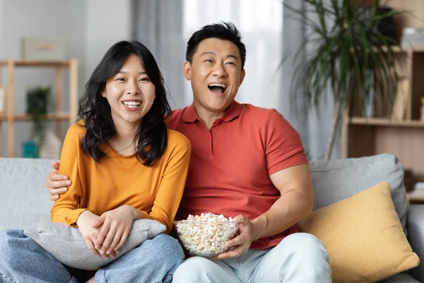 Cheerfy asian middle aged man and young woman in homewear sitting on sofa in living room, hugging, eating popcorn and laughing while watching TV together at home, copy space