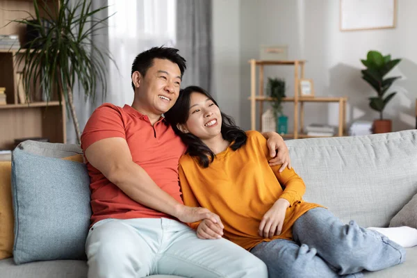 Loving asian middle aged husband and pretty young wife sitting on comfy couch at home, hugging and looking at copy space for advertisement, smiling, living room interior. Family, love, relationship