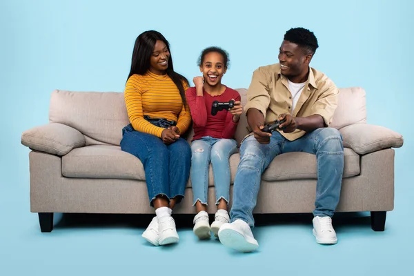 Gaming together. Playful african american father and daughter playing videogames and mother cheering while sitting on sofa over blue background. Parents and their child having fun
