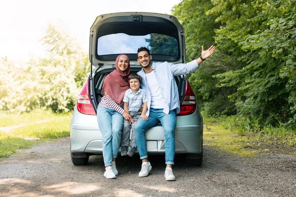 Positive auto journey. Happy millennial arabic guy and lady in hijab with baby, sit in car trunk and gesturing, resting from road. Family travel, vacation, relax and trip, human emotions, copy space