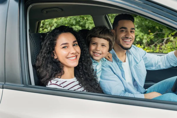 Cheerful glad millennial arab mother and father with kid go on vacation by auto and look at camera at open window. Family trip on new car, weekend together, love and relationship, positive emotions