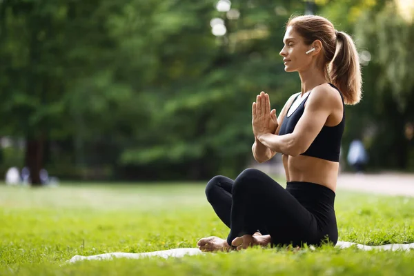 Peaceful millennial blonde woman in black tight sportswear sitting on fitness mat in lotus position with closed eyes, using wireless earbuds, meditating alone at public park, side view, copy space