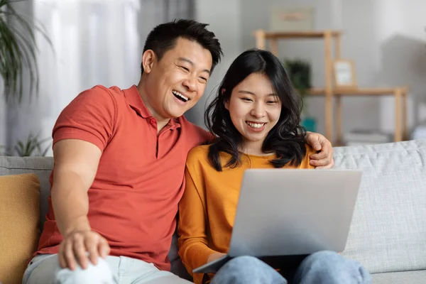 Happy chinese couple using laptop together, cozy home interior, loving asian man hugging his beautiful wife and looking at notebook screen, watching photos or funny content online, copy space