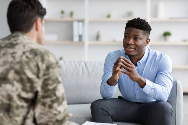 Handsome young black man professional psychotherapist meeting with veteran unrecognizable guy wearing camouflage uniform, helping soldier with reintegration after coming back home, clinic interior