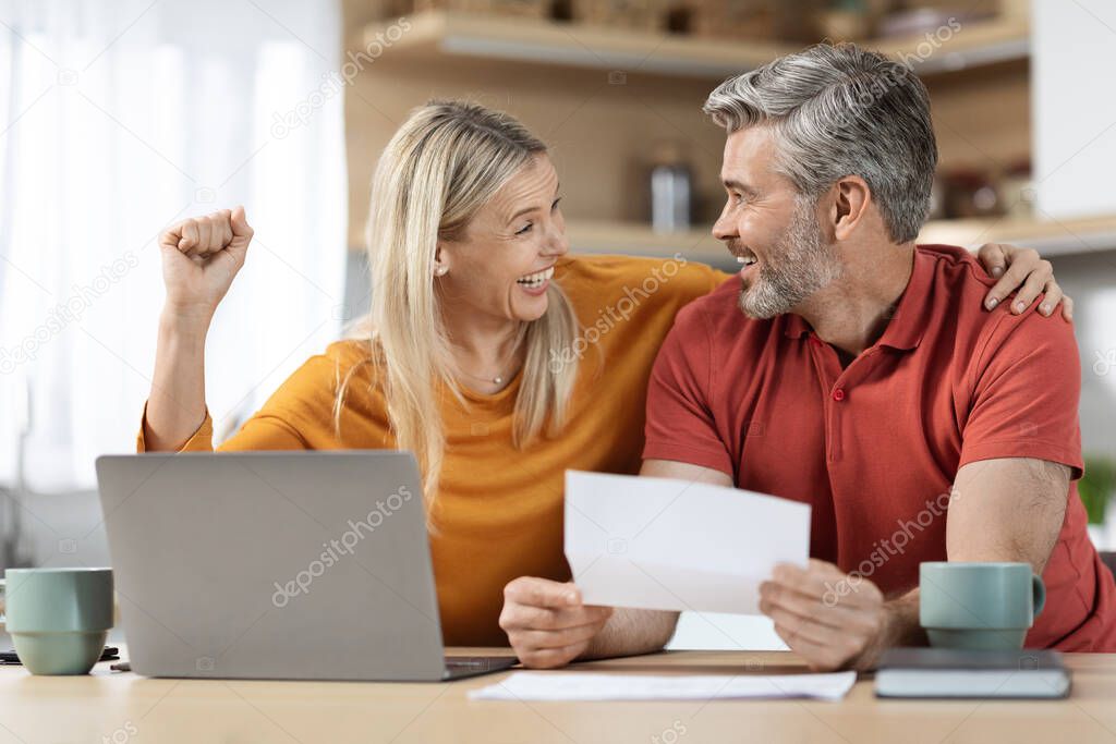 Emotional spouses sitting at table full of papers, laptop, coffee mugs, reading letter, smiling and raising hand up, got good news, kitchen interior. Economy, family budget, mortgage concept