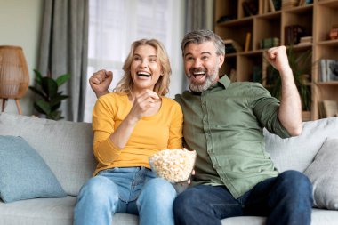 Weekend Pastime. Excited Middle Aged Couple Watching Sports On Tv At Home, Happy Mature Spouses Cheering Favorite Team And Eating Popcorn While Relaxing On Couch In Living Room Interior, Closeup Shot