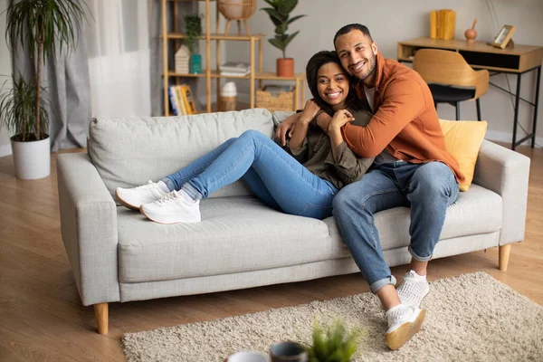 Romantic Relationship. Happy African American Spouses Hugging Smiling To Camera Sitting On Couch At Home. Husband Embracing Wife Resting On Sofa In Living Room. Marriage Concept