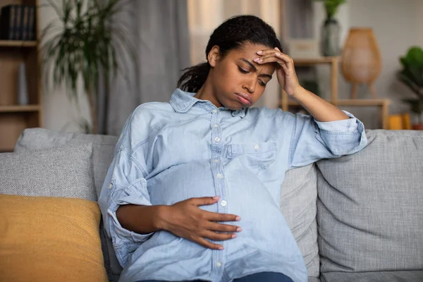 Pregnancy Sickness. Unhappy Pregnant African American Lady Feeling Bad Touching Head Having Headache Sitting On Couch At Home. Childbirth And Health Problems Concept