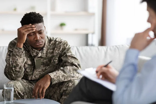 Upset african american man in military uniform veteran sitting on couch, touching his head while having conversation with psychologist, depressed soldier attending psychotherapist at clinic