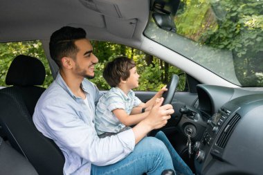 Happy smiling muslim young male teaches small kid how to drive, profile. Dad and son got drive lesson, new car, auto safety, life insurance, study at free time together. Love and family relationships