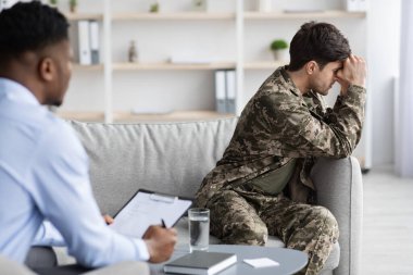 Upset young man in military uniform veteran sitting on couch, touching his head and crying while having conversation with black psychologist, depressed soldier attending psychotherapist at clinic