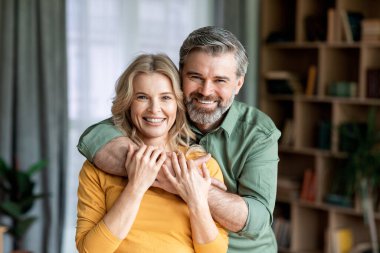 Portrait Of Loving Middle Aged Spouses Embracing And Smiling At Camera, Romantic Mature Couple Bonding While Posing In Living Room Interior, Enjoying Spending Time Together At Home, Copy Space