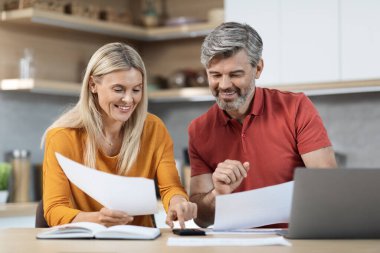Loving spouses attractive middle eastern man and woman sitting at kitchen table in front of computer, holding papers, using calculatir, working on family budget together, copy space