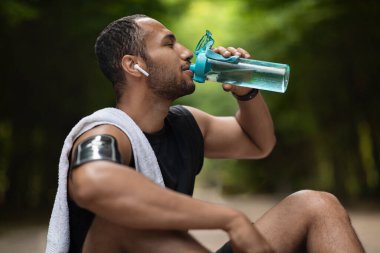 Athletic young black man drinking water while having break, training at public park, using wireless earbuds, listening to music while exercising, copy space. Hydration and sport concept