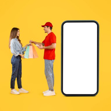 Shopping Delivery. Cheerful Deliveryman Giving Shopper Bags To Lady Near Big Smartphone With Empty Screen Over Yellow Studio Background. Square, Full Length
