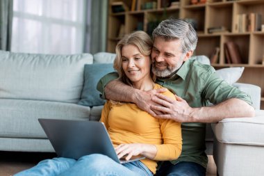 Portrait Of Romantic Middle Aged Spouses Relaxing With Laptop At Home, Happy Mature Couple Embracing And Using Computer, Resting On Floor In Living Room, Browsing Internet Or Shopping Online