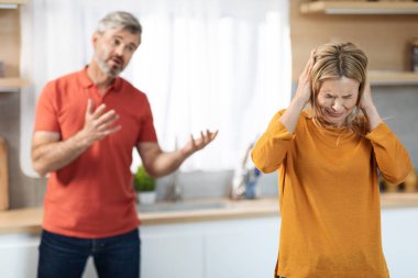 Stressful blonde woman crying while having quarrel with husband at home, touching her head, emotional middle aged man gesturing while talking to his upset spouse, kitchen interior