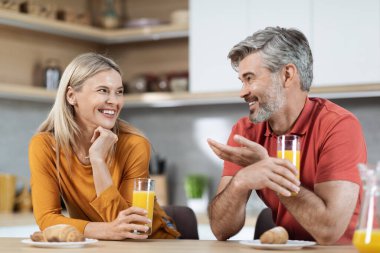 Happy loving spouses having fun while eating at kitchen, laughing and gesturing, cheerful loving husband and wife drinking fresh juice, eating fruits and pastry, having conversation, copy space