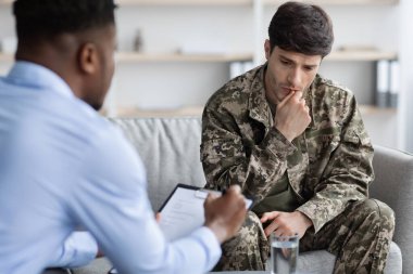 Pensive young man in military uniform soldier sitting on couch, touching his mouth and looking down while having conversation with black doctor, veteran attending psychotherapist at clinic
