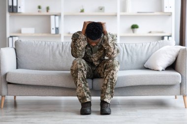 Stressed african american young man soldier in military uniform sitting on couch at psychotherapist office, covering his head and crying, having mental health issues, full length shot, copy space