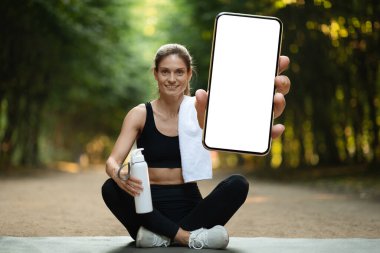 Positive young blonde woman in black sportswear sitting on fitness mat, holding bottle of water, doing yoga at park, showing smartphone with white empty screen, recommending app, mockup, copy space