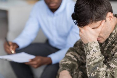 Top view of pensive young man in military uniform soldier sitting on couch, touching his head while having conversation with black doctor, veteran attending psychotherapist at clinic