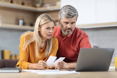 Shocked middle aged husband and wife sitting at kitchen table in front of laptop, counting spendings, checking bills, having finacnial problems, suffering from crisis while pandemic, copy space