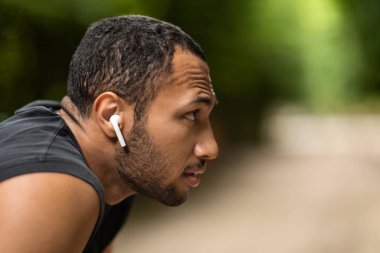 Profile portrait of attractive young black guy sportsman training outdoors, african american man enjoying music while jogging at public park, using wireless earbuds, looking at copy space