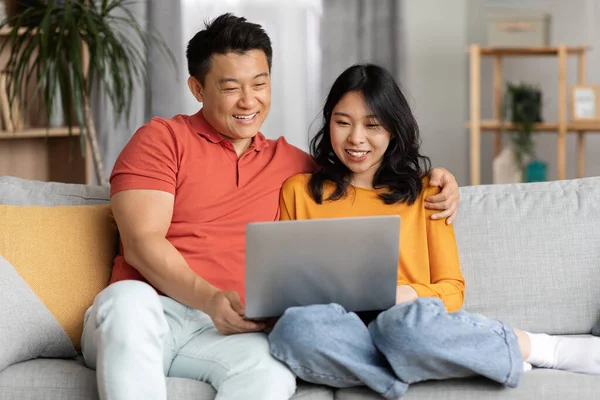 Asian lovers watching movie on Internet, using laptop at home. Smiling chinese man and woman sitting on couch, embracing, looking at computer screen, websurfing together, copy space