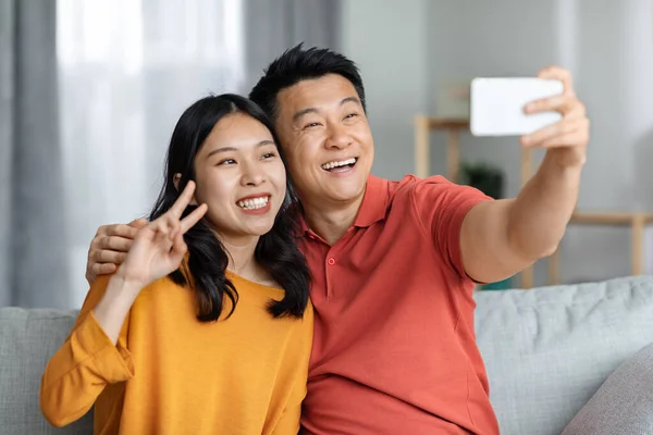 Loving asian couple using cell phone, sitting on couch in cozy living room, embracing, smiling, taking selfie, happy chinese man holding smartphone, pretty woman showing peace gesture, closeup