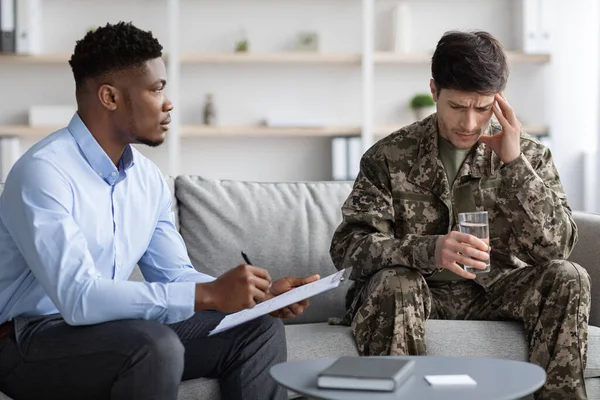 African american psychotherapist having session with soldier suffering from military trauma, psychologist asking veteran questions, taking notes, combat with glass of water in his hand touching head