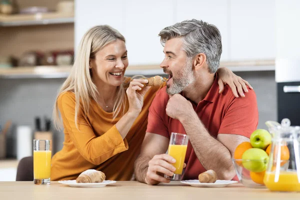 Positive blonde woman feeding her handsome spouse, attractive loving middle aged couple enjoying healthy delicious breakfast together, modern kitchen interior. Family, love, relationships concept