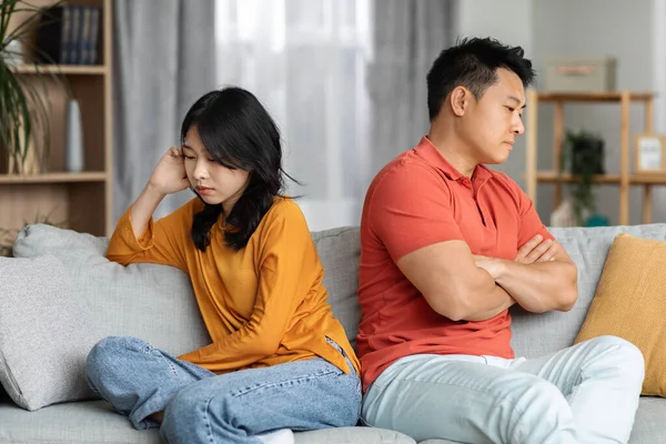 Portrait of frustrated asian spouses after fight, upset man and woman sitting on couch far from each other, looking down, thinking about divorce, breakup after quarrel, home interior