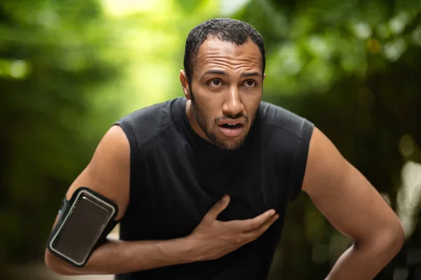 Handsome young black sportsman with heart pain touching his chest, jogging in the public park, using armband with smartphone, copy space. Sport and cardiovascular diseases concept