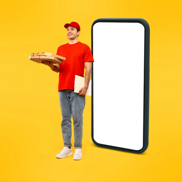 Food Delivery App. Cheerful Deliveryman Holding Pizza Boxes Standing Near Huge Smartphone With Blank Screen Posing Wearing Red Uniform And Cap On Yellow Background. Courier Service. Square