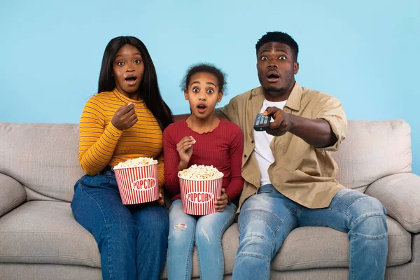 Young shocked black parents and their daughter watching movie on TV and eating popcorn, man pointing at camera with remote control, sitting on couch over blue studio background