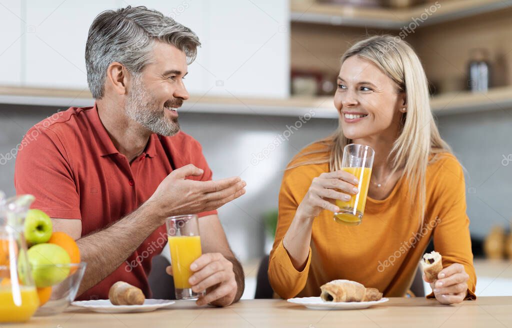 Beautiful romantic couple having breakfast together at home, cheerful middle aged man and woman in casual outfits sitting at kitchen table, drinking juice, eating pastry, talking and smiling
