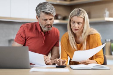 Upset middle aged husband and wife sitting at table in front of computer, reading letter from bank, shocked with their debts, kitchen interior, copy space. Financial hangover during 2022 crisis