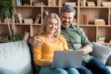 Smiling Middle Aged Spouses Resting With Laptop In Living Room Interior, Happy Mature Spouses Embracing And Using Computer While Relaxing Together At Home, Browsing Internet Or Shopping Online