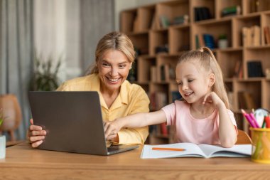 Happy teacher having class with cute little girl, doing homework together using laptop computer, sitting at home interior. Tutoring, private lessons, e-learning concept